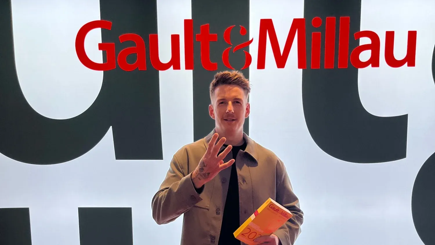 Clemens Gesser holding up four fingers for his four toques in front of the Gault&Millau logo, with the 2027 guide in his hand