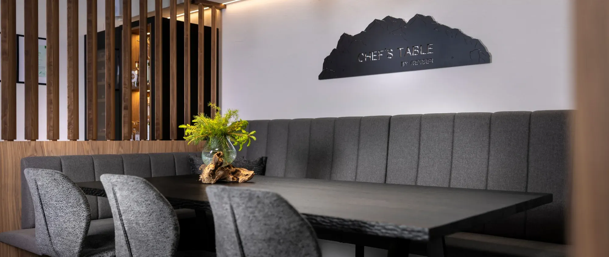 The Chef's Table at Hotel Gesser, a long dark table with grey chairs in front of the kitchen