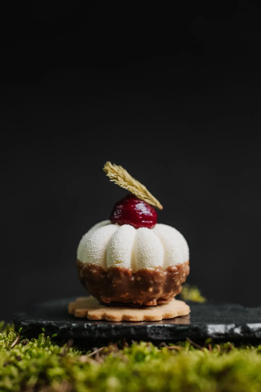 A white dessert topped with a glazed cherry on dark slate and moss at Chef's Table by Gesser