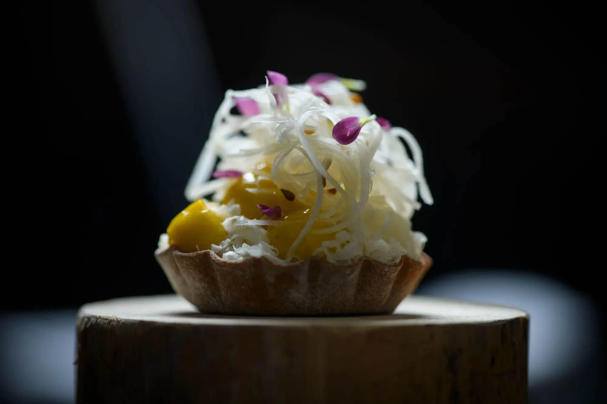 A tartlet topped with fine strips and edible flowers on a wooden stump at Chef's Table by Gesser
