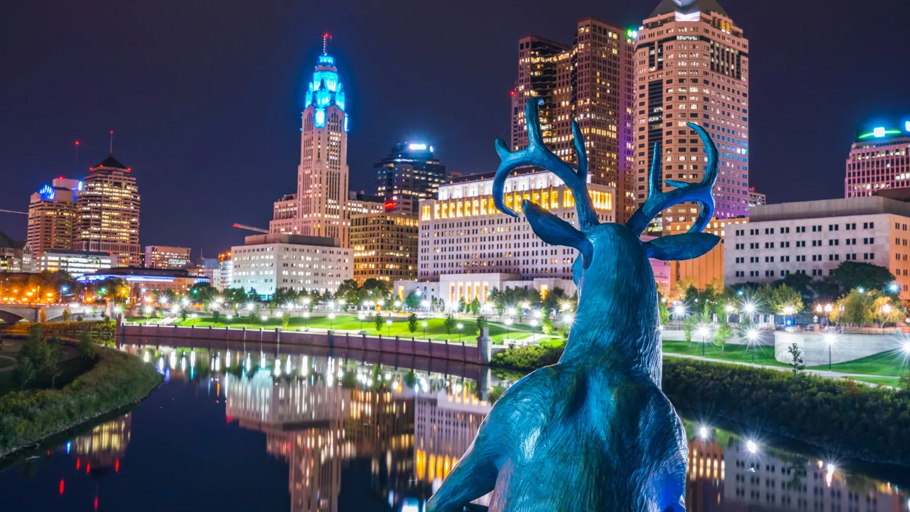 The skyline of Columbus, Ohio, at night, reflected in the Scioto River, with a bronze stag statue in the foreground