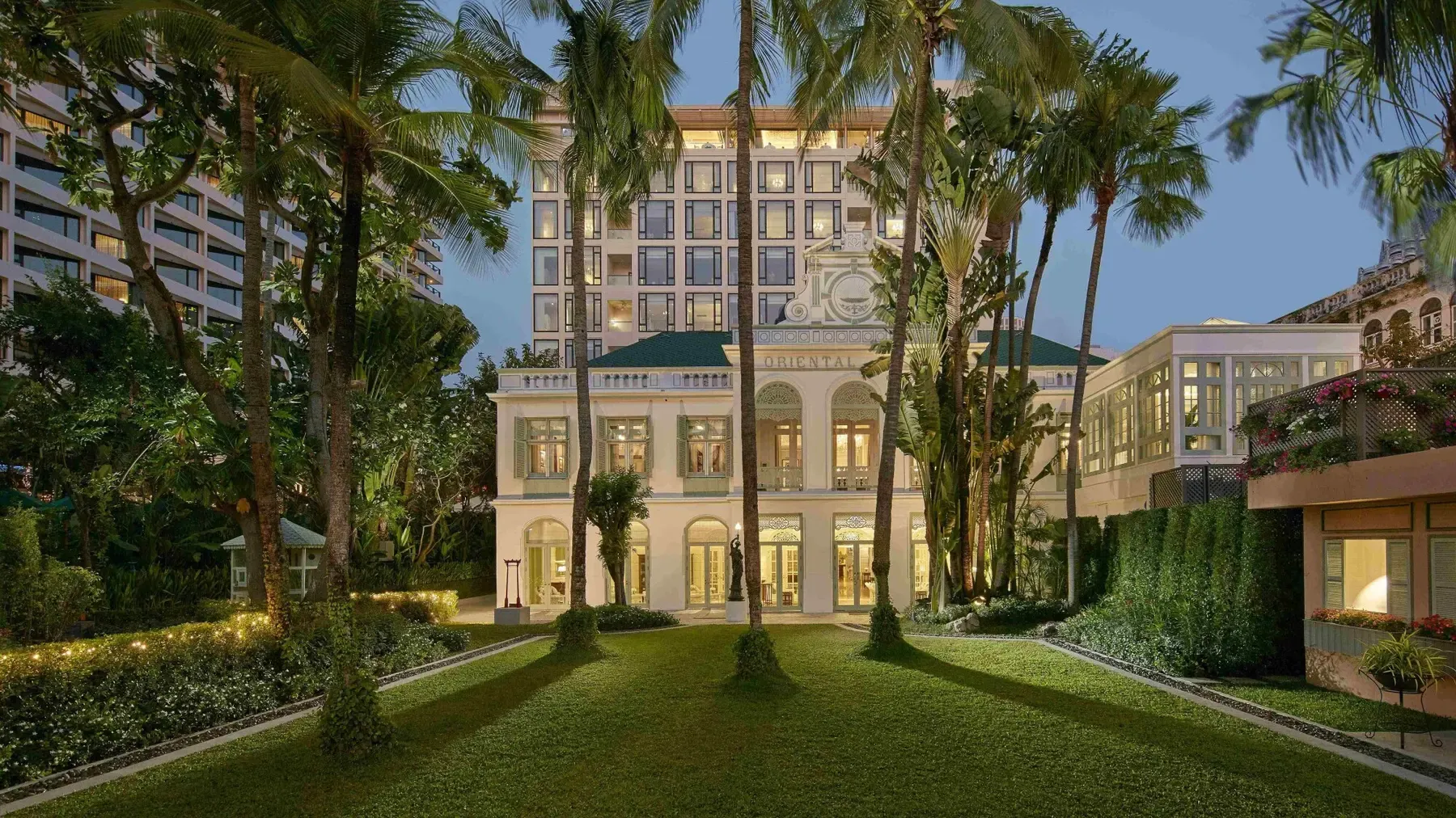 A white colonial-style Mandarin Oriental hotel building with a lawn and tall palm trees at dusk